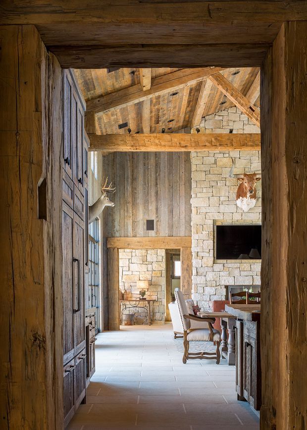A rustic, lodge-style interior viewed through a substantial timber doorway. The space features exposed dark timber beams and timber wall and ceiling lining, pale stone masonry walls, a large stone fireplace, tall vertically proportioned volumes, and a pale stone or tile floor. Traditional upholstered dining chairs and a timber dining table are visible in the foreground, with a secondary sitting area beyond. Mounted antlers and animal heads reinforce the alpine hunting-lodge character.