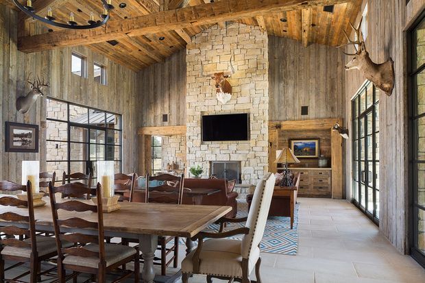 A spacious rustic great room combining dining and living areas, with exposed timber ceiling beams and wall cladding, a large pale stone fireplace surround with integrated television, extensive black-framed glazing, and substantial timber dining furniture. Mounted hunting trophies and leather details add a lodge-inspired character, while a patterned rug and upholstered dining chairs soften the room.