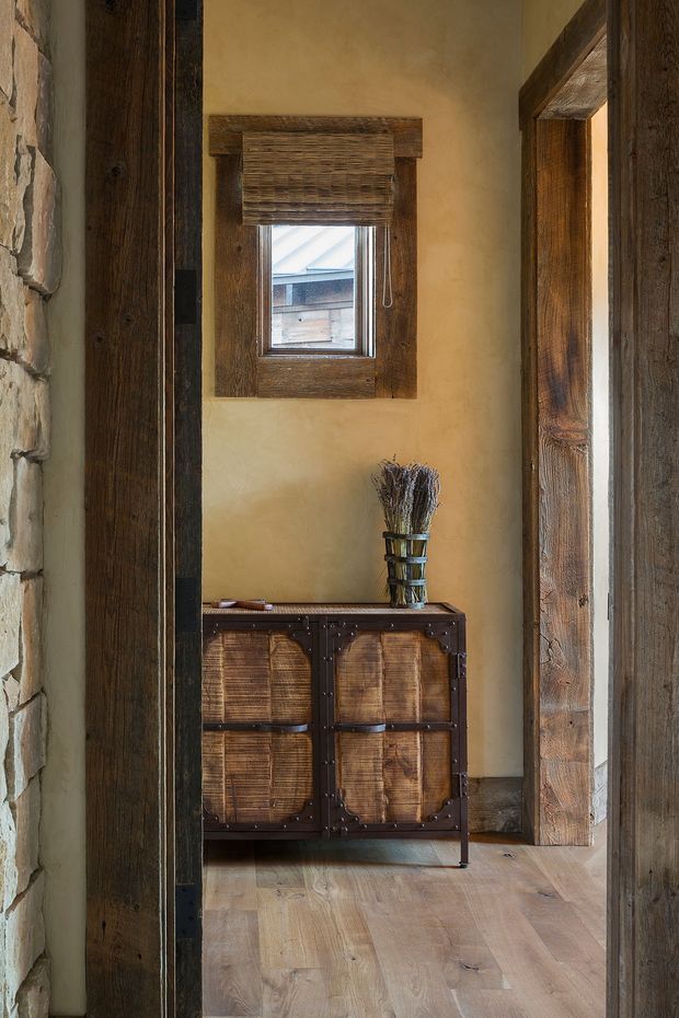 A narrow rustic entry hall framed by heavy reclaimed timber posts and exposed stonework. The warm plaster walls surround a small window with a woven Roman blind, while a dark wood and woven-rattan console cabinet sits beneath it. Wide timber floorboards and substantial timber door frames reinforce the earthy, handcrafted character.
