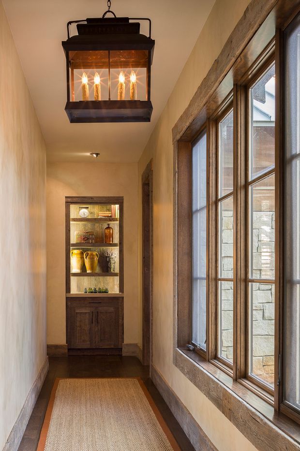 A narrow, warmly lit residential hallway with a dark timber floor, beige plaster walls, and a long run of timber-framed windows on the right. A rustic black metal lantern pendant hangs from the ceiling, while a built-in dark timber display cabinet with open shelves and lower cupboards anchors the far end. The space has a traditional, rustic character with natural timber detailing and warm ambient lighting.