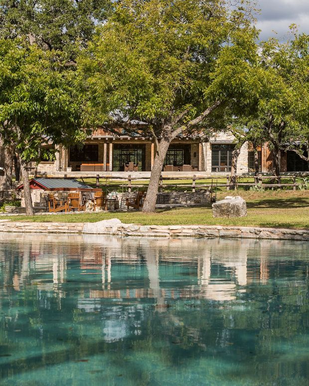 A low-rise stone-clad building viewed across a clear body of water, set within a mature landscaped garden. The facade features broad dark-framed glazed openings, stone columns and walls, and a covered outdoor seating area with timber furniture. Trees partially screen the building, while the water dominates the foreground and reflects the architecture and vegetation. The setting has a rustic, resort-like character.