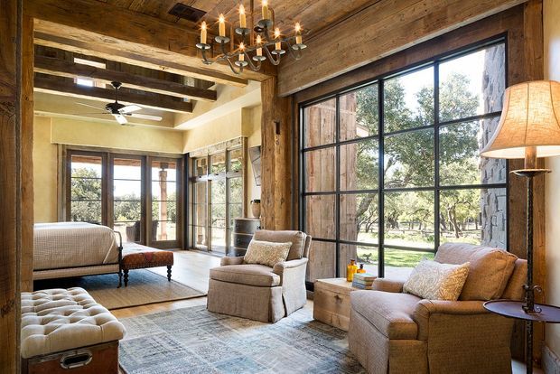 A warm, rustic living room with large black-framed windows, exposed timber ceiling boards and beams, timber-clad walls, and a sitting area arranged around an upholstered armchair and sofa. A bedroom or sleeping area is visible through the open framed connection at the rear, while a chandelier, table lamp, ottoman, and patterned rug add layered traditional character.