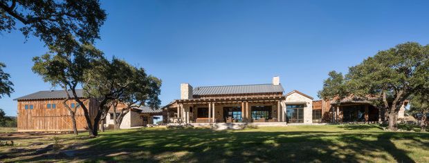 Wide exterior view of a low, sprawling residential property set across a landscaped lawn. The main building has a pitched metal roof, stone-clad chimney forms, timber-framed glazing and a covered veranda, with timber outbuildings or ancillary structures extending to both sides. Mature trees frame the composition under a clear blue sky.