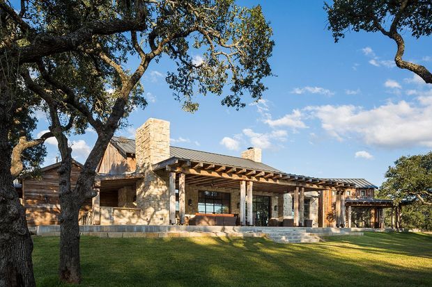 A broad exterior view of a low-slung stone-and-timber residence set among mature oak trees. The house has a long metal-clad pitched roof, prominent masonry chimneys, timber cladding, extensive glazing, and a deep timber pergola/verandah over the main entrance and terrace. A manicured lawn and strong afternoon shadows frame the building against a clear blue sky.