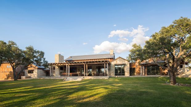 A single-storey low-rise residence viewed from a broad landscaped lawn. The home has a long, informal composition with a pitched corrugated metal roof, exposed timber porch structure, stone chimney and substantial stone masonry walls, plus large glazed openings and warm timber cladding. Mature trees frame the property and the image has a sunny, relaxed rural or suburban character.