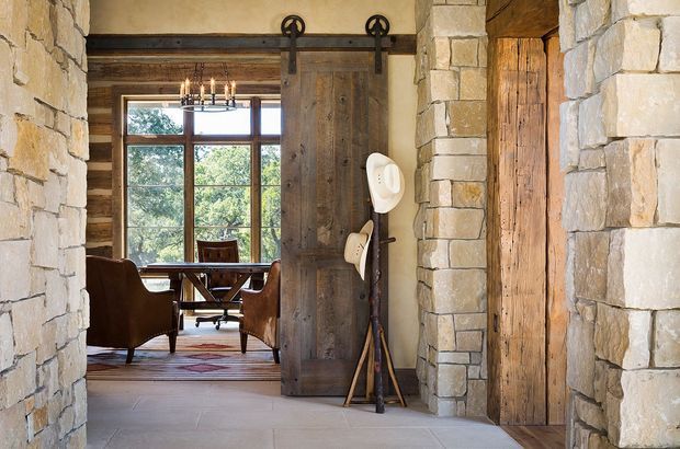 Rustic stone-lined interior entry or passage with a large weathered timber barn-style sliding door, opening into a warm dining or living area. The adjoining room features exposed timber beams, expansive framed glazing, a chandelier, wooden dining furniture, and leather armchairs. Natural stone walls and timber finishes create a lodge-like, traditional atmosphere.