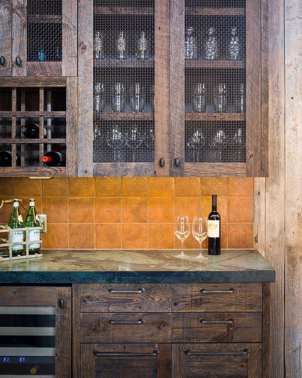 Close-up of a rustic built-in home bar or wine storage area. Dark weathered timber cabinetry with wire-mesh glass-front doors stores wine glasses and bottles above a patterned terracotta tile splashback. A dark stone benchtop sits over timber drawers and cabinetry, with a wine fridge visible below.