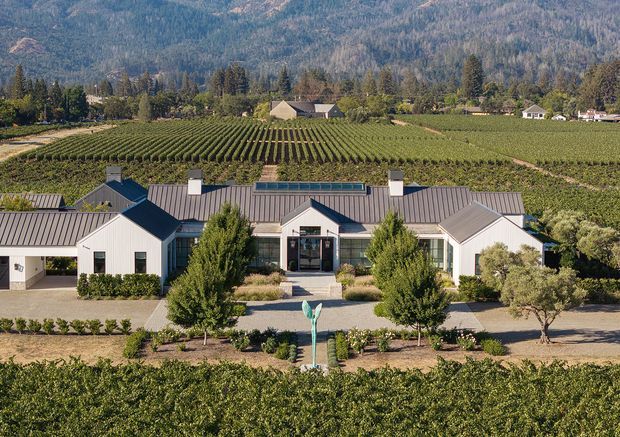 A low, symmetrical rural estate building set amid orderly vineyard rows, with white gabled wings, dark standing-seam metal roofs, a glazed central entry, landscaped frontage, and forested mountains in the background.