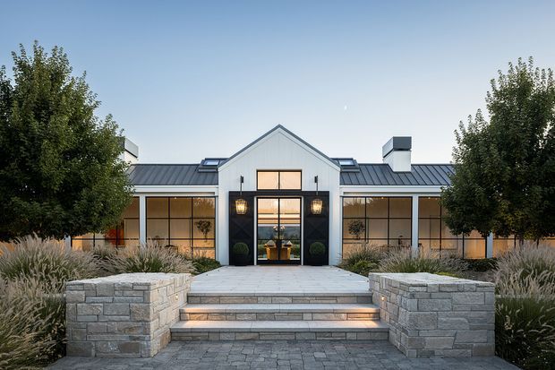 Symmetrical single-storey contemporary house facade viewed from the landscaped front approach. The building features a central gabled entry pavilion with tall glazed doors and windows, flanking flat-roofed or low-pitched wings, dark standing-seam metal roofing, pale vertical cladding, warm interior lighting, and broad stone steps bordered by substantial rendered or masonry planter walls. Mature trees and ornamental grasses frame the entrance.