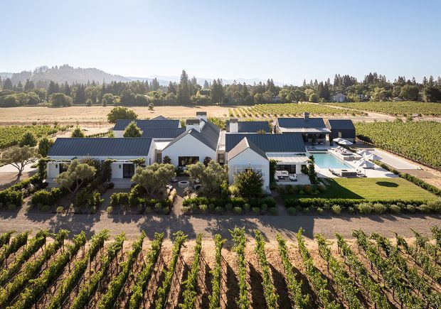 Aerial view of a large contemporary rural estate surrounded by orderly vineyard rows and cultivated farmland. The property consists of several connected or clustered white-rendered buildings with dark pitched metal roofs, landscaped gardens, outdoor seating, and a swimming pool. Tree-covered hills and additional agricultural land form the background.