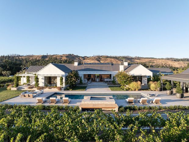 A wide exterior view of a low, single-storey residence arranged around a rectangular swimming pool and paved entertaining area. The home has light-coloured rendered walls, dark standing-seam metal gabled roofs, extensive glazing, landscaped gardens, and a vine-covered foreground. Dry rolling hills form the distant backdrop.