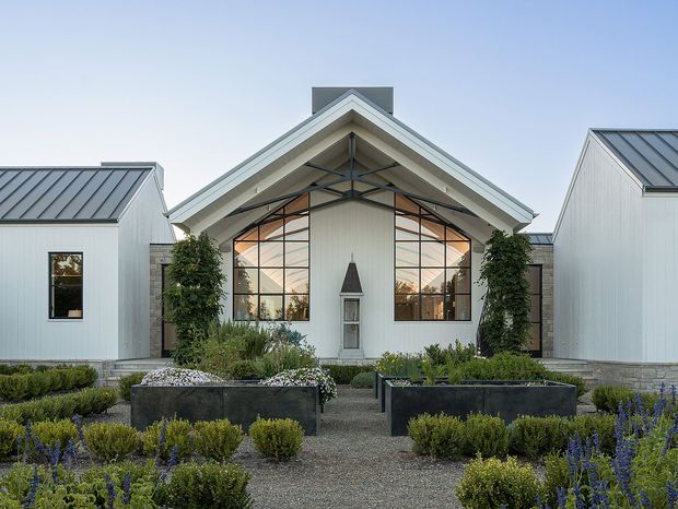 Front exterior view of a contemporary gabled building composed of three connected white-clad volumes. The central pavilion has a prominent pitched roof, exposed dark structural framing, and expansive glazed gable windows, flanked by smaller white wings with standing-seam metal roofs. A formal landscaped forecourt features clipped shrubs, raised dark planter boxes, gravel paths, and flowering plantings.