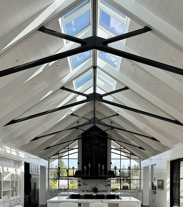 A dramatic, bright kitchen interior in a high vaulted, gabled space. White wall panelling and cabinetry frame a central kitchen island, while exposed black steel trusses span beneath a glazed roof ridge with multiple skylights. Large black-framed windows overlook trees and bring in abundant daylight. A dark central feature structure and suspended linear chandelier create strong contrast against the white interior.