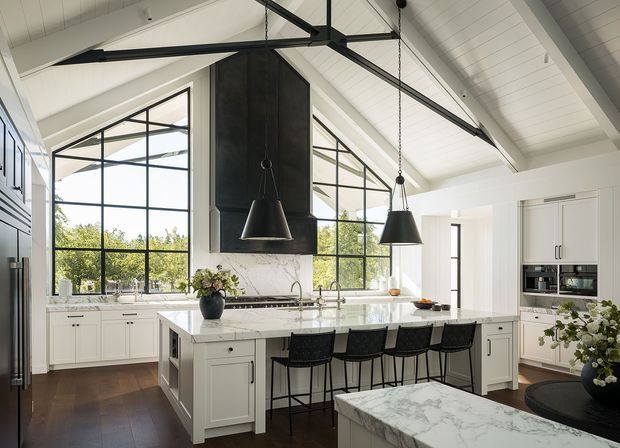Bright, spacious kitchen with a large marble-topped island, white shaker-style cabinetry, integrated appliances, black-framed steel-look glazing, and dramatic black pendant lights. The room has a high vaulted white-painted ceiling with exposed dark structural beams, extensive windows overlooking greenery, and dark timber flooring. A marble-clad rangehood forms a strong central feature above the cooktop, while a secondary marble-topped surface and seating occupy the foreground.