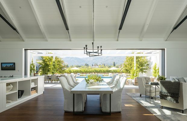 Bright, expansive dining and living area with a long dining table, upholstered dining chairs, and a contemporary chandelier beneath a white beamed ceiling. Wide open glazing frames a pool, landscaped garden, and distant mountain scenery. A built-in white shelving unit and seating area occupy the sides of the room, while dark timber flooring adds contrast to the light interior.