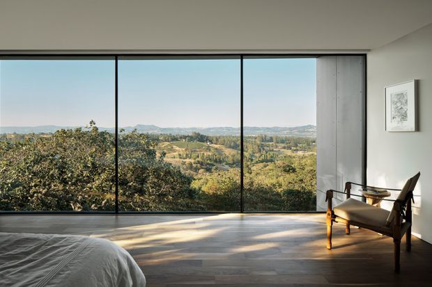 A serene contemporary bedroom with a low, uncluttered profile and expansive floor-to-ceiling glazing overlooking a wooded rural landscape and distant hills. The room has pale smooth walls, dark timber flooring, a partially visible bed with white bedding, and a sculptural timber-framed lounge chair beside the window. Strong natural light creates long shadows across the floor.