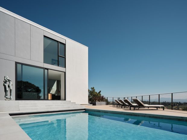 A minimalist two-storey flat-roof house with a pale concrete or rendered facade, large dark-framed floor-to-ceiling windows, and broad steps leading to a rectangular swimming pool. Poolside loungers sit on a paved terrace overlooking a distant landscape under a clear blue sky.