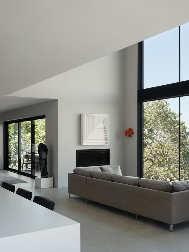A light-filled contemporary open-plan living and dining space with a large grey sectional sofa facing a sleek built-in black fireplace. White walls and ceilings, pale grey floor tiles, expansive black-framed floor-to-ceiling glazing, and minimalist sculptural artwork create a clean, refined interior. A white dining table with black chairs occupies the foreground, while leafy trees are visible outside.