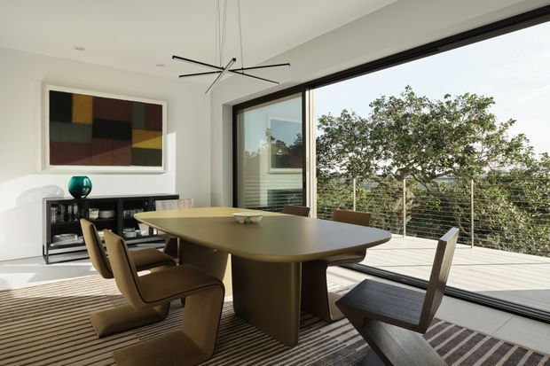 A bright, refined dining room with a large sculptural oval dining table and contemporary dining chairs. Full-height black-framed glazing opens onto a timber deck, while a low dark sideboard, abstract artwork and pendant-style ceiling fan add visual interest. The room has white walls, a striped area rug and a leafy outdoor outlook.