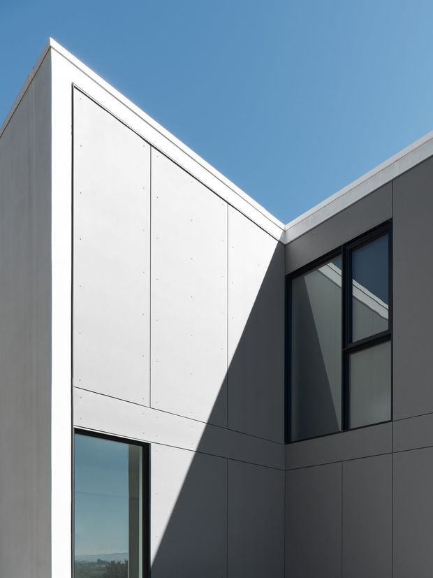 Close-up exterior architectural photograph of a sharply angular, minimalist building facade under a clear blue sky. The composition shows pale smooth panel cladding, dark recessed wall sections, crisp rooflines forming a strong V-shaped junction, and tall dark-framed windows with reflective glazing. Strong sunlight creates a dramatic triangular shadow across the facade.