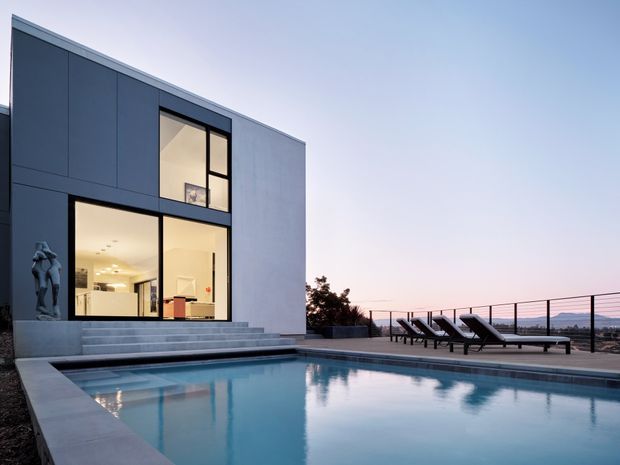 Contemporary flat-roofed residence viewed from the pool terrace at dusk. The dark-panelled and pale rendered facade has expansive floor-to-ceiling glazing opening to a brightly lit interior, with broad entry steps leading down to a rectangular swimming pool. Minimal poolside loungers, a slim metal balustrade, and distant landscape create a clean, elevated modern setting.