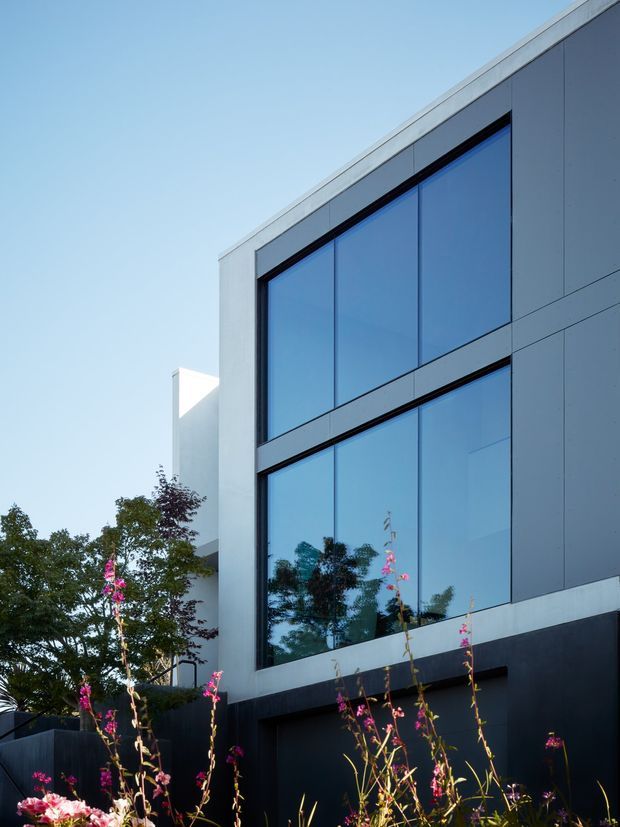 A close-up exterior architectural view of a contemporary multi-storey building. The facade combines large blue-tinted glazed window panels with dark horizontal framing, pale grey concrete or panel surrounds, and darker lower wall cladding. A clean flat roofline and strong geometric composition dominate the image, with landscaped flowering plants in the foreground.