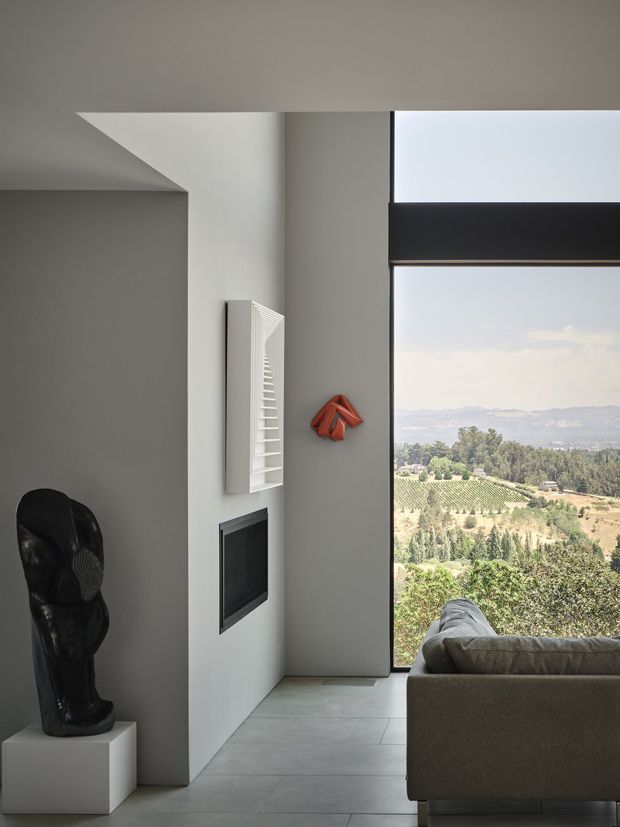 A refined minimalist living room with a sculptural black artwork on a white plinth, a wall-mounted linear fireplace or dark inset feature, white louvered wall detail, and a grey upholstered sofa. Floor-to-ceiling glazing frames a rural vineyard and wooded landscape. The space has crisp geometric forms, pale grey tiled flooring, neutral walls, and strong natural light.