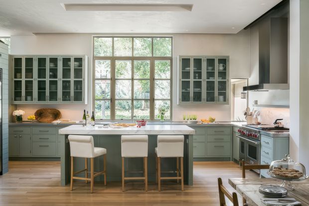 A bright, spacious kitchen with a large central island, pale sage-green cabinetry, glass-front upper cabinets, white countertops, timber flooring, and a substantial multi-pane window. The island includes seating for three, while a professional-style range and large stainless-steel rangehood occupy the right side. A dining table is partially visible in the foreground.