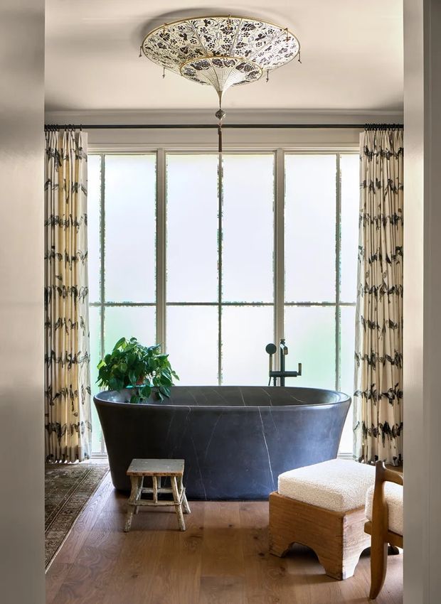 Elegant bathroom featuring a freestanding dark charcoal bathtub positioned in front of tall multi-pane frosted windows, framed by patterned floor-length curtains. A decorative ornate pendant light hangs above the tub, with a small timber stool, upholstered bench, indoor plant, timber flooring, and dark metal bathtub tapware completing the room.