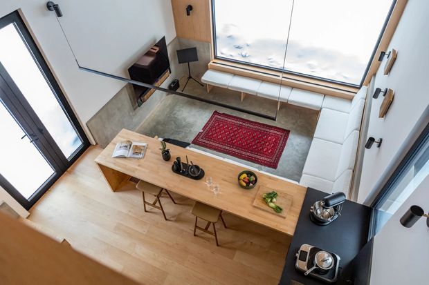 High-angle view of a double-height contemporary living and dining area. A long timber dining island with stools occupies the foreground, while a white sectional sofa, red patterned rug, fireplace, and large skylight-style glazing define the lounge area. Black-framed glazing, pale timber flooring, concrete-effect wall surfaces, and black kitchen or bar cabinetry create a refined modern interior.