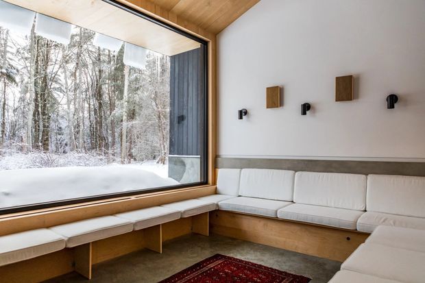 A bright, minimalist lounge with a built-in U-shaped upholstered window seat and sectional-style seating surrounding a low table area. Full-height corner glazing overlooks a snowy forest, while warm timber lines the ceiling, window frame, and built-in seating base. The white walls feature simple black wall lights and small timber wall accents, creating a calm contemporary retreat.