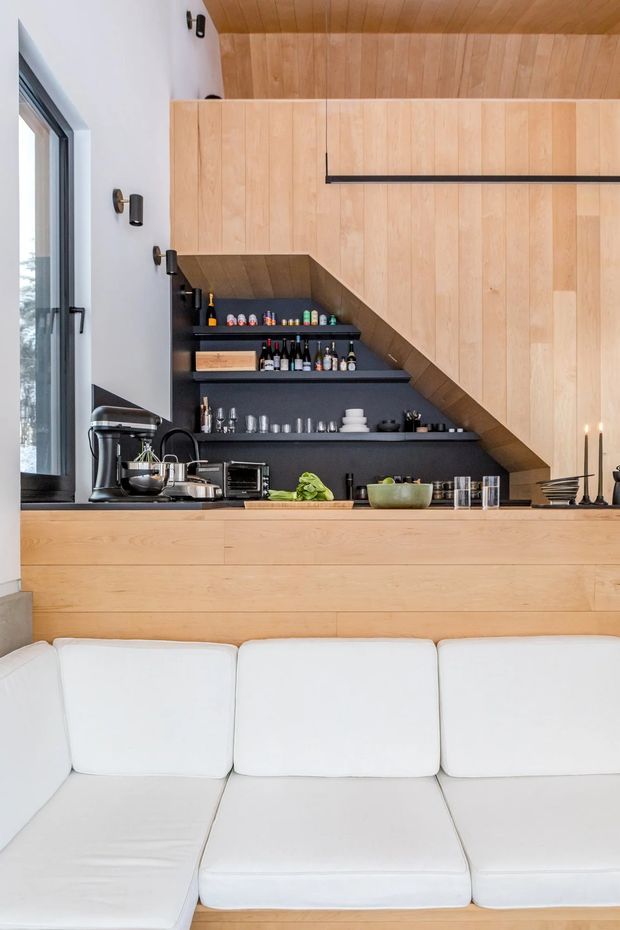 A bright, compact open-plan living area featuring a white sectional sofa in the foreground and a timber-clad kitchen or entertaining bar behind it. The kitchen includes dark open shelving stocked with glassware and bottles, a countertop appliance, and light timber cabinetry. A dramatic sloped timber ceiling and matching wall panels create a warm, contemporary built-in interior, with black-framed glazing and linear pendant lighting providing contrast.