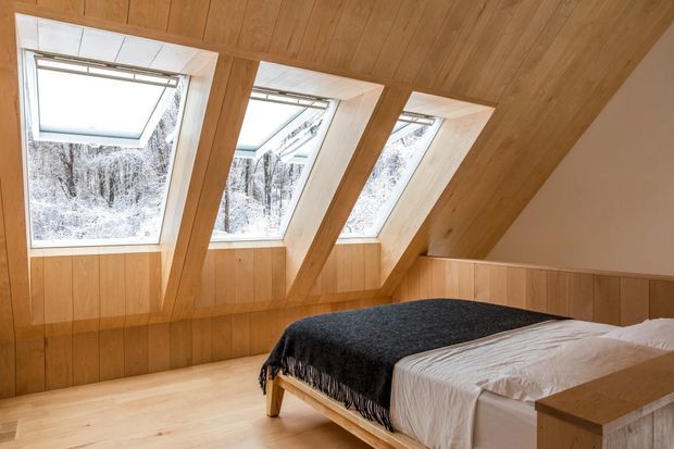 A warm attic bedroom beneath a sloping timber-clad roof, featuring three large angled skylights, timber wall panelling, pale timber flooring, and a simple bed with a dark throw. Snow-covered trees are visible through the glazing, and the space has a bright, calm, natural character.