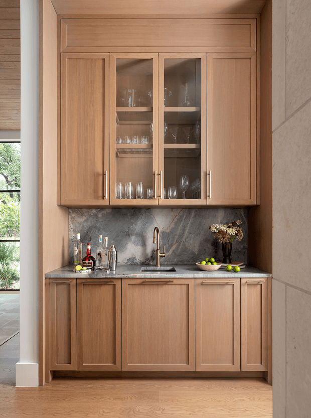 A compact, built-in drinks bar with warm timber shaker-style cabinetry, upper glass-front cupboards displaying stemware, a dark veined stone backsplash and benchtop, and a small undermount sink with brass-toned tap. Bottles, glasses, citrus, and a vase provide restrained styling. The joinery is set into a light-toned architectural niche beside a large window, with timber flooring continuing through the space.