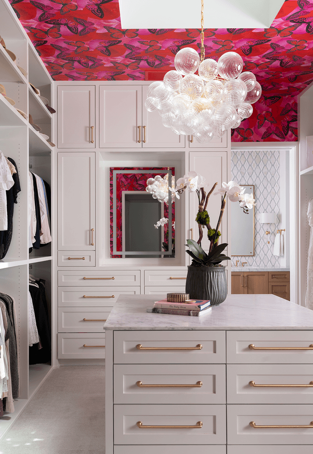 A bright, highly styled walk-in wardrobe and dressing room with extensive pale built-in cabinetry, open clothing shelving, drawers, and a central island with a light stone benchtop. The room features a vivid pink and red butterfly-patterned ceiling and matching mirror surround, gold-toned drawer pulls, a sculptural clear-glass bubble chandelier, and a patterned light wall panel with a small timber vanity area. An orchid arrangement sits on the island.