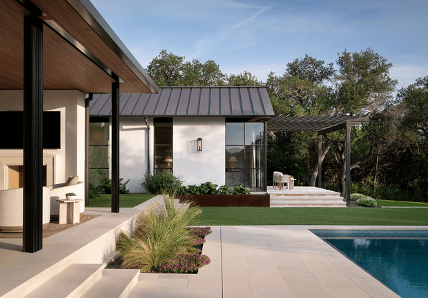 Contemporary single-storey residence viewed from a landscaped outdoor terrace, with crisp white rendered walls, dark metal pitched roofing, timber-lined soffits, slender black structural posts, and expansive glazing. A paved pool terrace occupies the foreground, while a covered outdoor dining area, raised planter beds, ornamental grasses, and mature trees create a refined connection between the house and garden.
