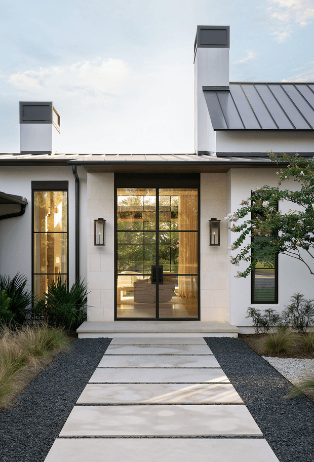 Front exterior view of a contemporary single-storey residence. The white rendered facade has a strong central glazed entry, black-framed windows and doors, slim wall-mounted lanterns, and a standing-seam metal roof with two tall chimneys. A minimalist landscape uses large pale concrete stepping slabs set into dark gravel, with ornamental grasses and sculptural planting.