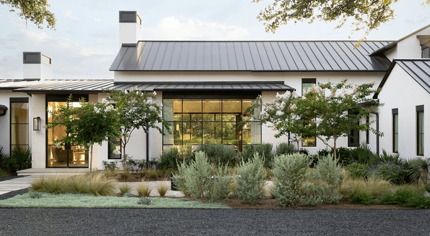 A single-storey contemporary residential building viewed from the landscaped front garden. The white rendered facade is composed of connected wings beneath dark grey standing-seam pitched metal roofs, with tall narrow windows, large black-framed glazed openings, and prominent white chimneys. A layered drought-tolerant landscape with ornamental grasses, silvery shrubs, and small trees forms the foreground.