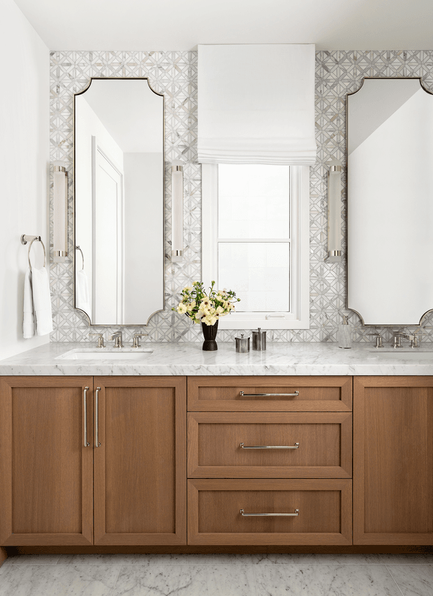 A bright, refined bathroom vanity with a symmetrical double-sink arrangement. Warm wood cabinetry with recessed-panel doors and drawers supports a white marble countertop, while two ornate framed mirrors flank a central window dressed with a white Roman shade. The backsplash is covered in a geometric patterned tile, with polished metal plumbing fixtures, wall sconces, and a small floral arrangement adding detail.