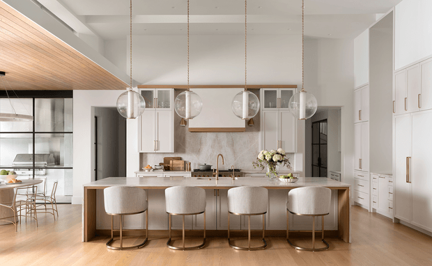 A bright, refined open-plan kitchen with a large island and four upholstered bar stools. The room features white shaker-style cabinetry, a pale stone benchtop and splashback, warm timber flooring, a timber-lined ceiling section, black-framed glazed partitions, and four spherical glass pendant lights. A dining area is visible to the left, while tall built-in storage occupies the right wall.