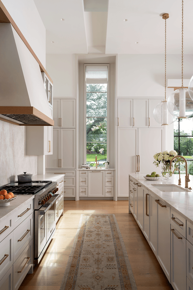 Bright, refined kitchen interior with a long central island, extensive white panelled cabinetry, integrated window-seat or sink-wall storage, and a professional range beneath a sculptural hood. A tall window brings in garden views and natural light. Brass-toned hardware, a pale stone benchtop, timber flooring, and globe pendant lights create a warm traditional-contemporary feel.