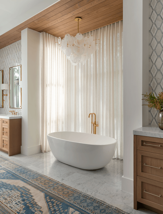 Bright, refined bathroom featuring a freestanding white oval bathtub in front of sheer floor-to-ceiling curtains, with a gold-toned floor-mounted faucet. Warm timber plank ceiling, patterned geometric wallpaper, marble-look flooring and benchtops, and timber bathroom vanities create a polished transitional aesthetic. A large blue patterned rug and a sculptural glass pendant add decorative character.