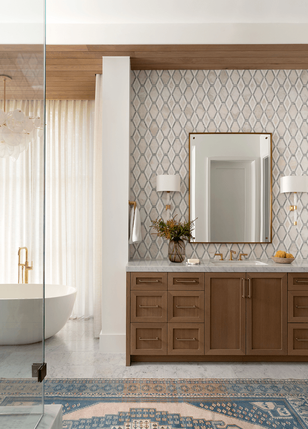 A refined, spacious bathroom featuring a freestanding white bathtub behind a frameless glass shower panel, a large timber vanity with brass hardware, pale stone benchtop, patterned geometric wall tile, and a substantial framed mirror. White curtains diffuse daylight, while a blue patterned rug and warm timber cabinetry add contrast and softness.