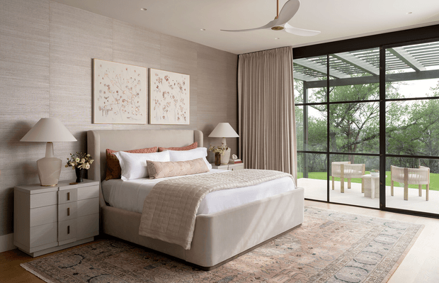 A refined contemporary bedroom with a softly upholstered bed, layered white and beige bedding, matching bedside tables and lamps, delicate botanical artwork, textured neutral wall covering, full-height curtains, and expansive black-framed glazing opening toward a green outdoor setting. A large patterned rug and sculptural ceiling fan add warmth and detail.