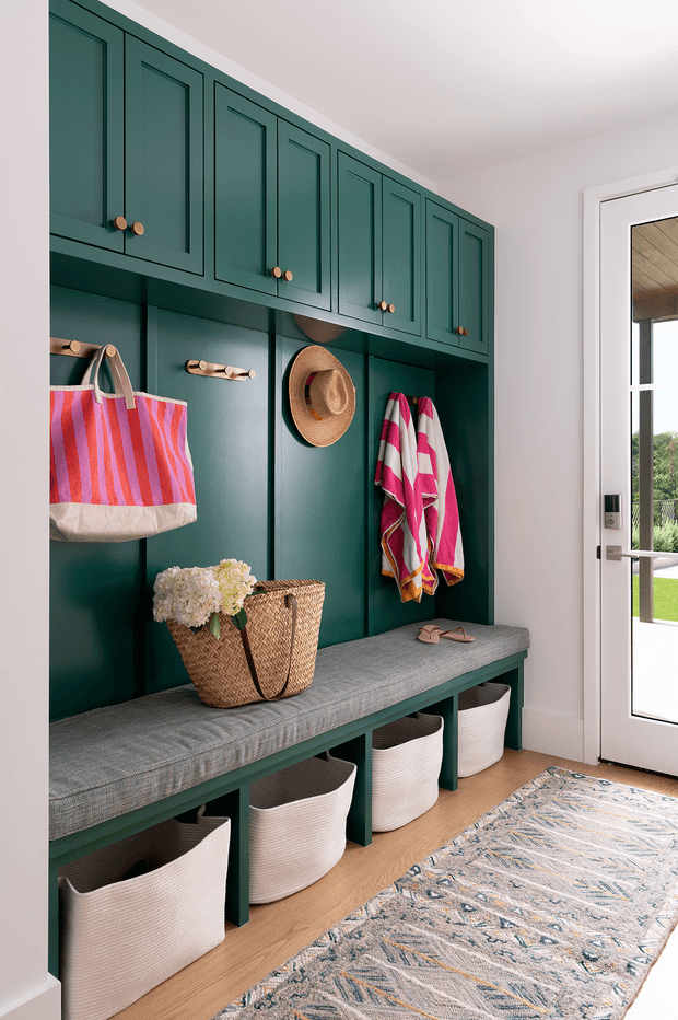 A bright, neatly organised mudroom or entryway with deep green built-in cabinetry and open cubby storage. The joinery includes framed upper cabinet doors with small round wood knobs, a continuous upholstered bench seat, hooks holding bags and towels, woven baskets below, and a textured patterned runner on light timber flooring. A glazed exterior door provides natural light and a view to the outdoors.