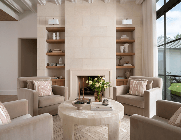 Bright, refined living room centered on a round stone coffee table and fireplace. Four upholstered armchairs surround the table, while symmetrical built-in timber shelving and cabinetry flank a pale stone fireplace surround. Tall glazing brings in abundant daylight, and exposed painted ceiling beams add architectural character.