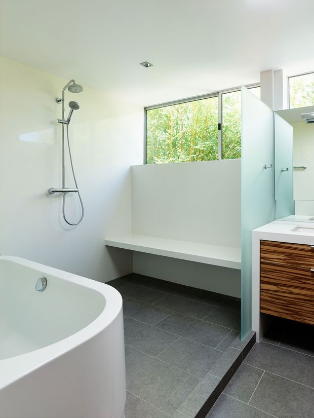 Bright contemporary bathroom with a freestanding white bathtub in the foreground, open walk-in shower with dual showerheads, a built-in bench, grey tiled flooring, large frosted or translucent windows, and a floating timber vanity with a white benchtop. The space has clean lines and abundant natural light.