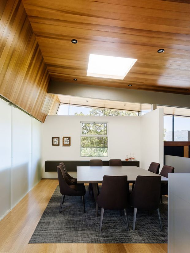 Bright contemporary dining room with a large rectangular dining table and dark upholstered chairs arranged on a textured rug. The room features extensive warm timber lining across the angled ceiling and upper wall, white walls, a long dark built-in sideboard, clerestory glazing, a skylight, and views through the windows.