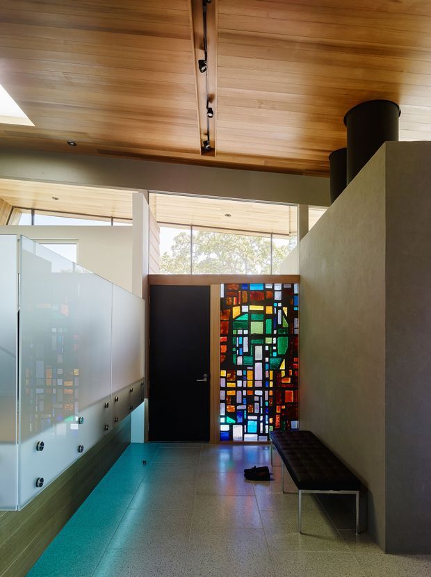 A bright, architecturally detailed interior hallway or entry space with a warm timber-lined ceiling, clerestory glazing, polished terrazzo-like flooring, frosted glass screening, a dark built-in or fixed bench, and a vivid geometric stained-glass feature beside a dark door. The space has strong mid-century modern character, with clean lines and abundant natural light.