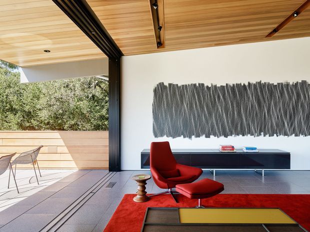 A refined modern living room opening onto a timber-clad outdoor terrace. The space features a warm timber-lined ceiling with exposed dark structural beams and track lighting, pale walls, large-format grey flooring, a low dark built-in media cabinet, and a sculptural red lounge chair with matching ottoman on a bold red rug. A large monochromatic textured wall artwork provides a dramatic focal point, while expansive open glazing connects the room to the landscaped exterior.
